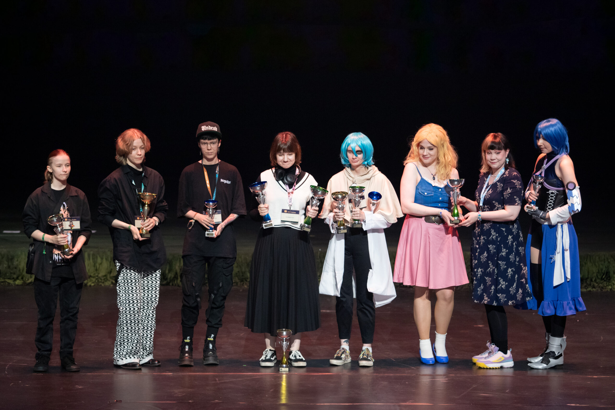 Vuoden 2024 AMV-kilpailun voittajat Tampere-talon Ison salin lavalla. Voittajat pitelevät pokaaleja kädessään ja näyttävät tyytyväisiltä. Joillain kilpailijoista on useampia pokaaleja.